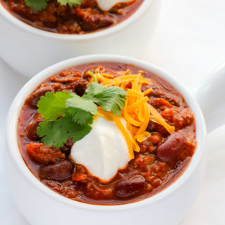 Overhead view of two bowls of Ground Beef Chili, garnished with shredded cheese, cilantro and sour cream, on a white surface.