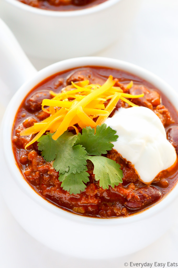 Close-up overhead view of a bowl of Ground Beef Chili, garnished with shredded cheese, cilantro and sour cream, on a white surface.