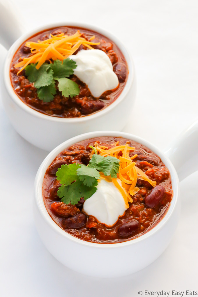 Overhead view of two bowls of Ground Beef Chili, garnished with shredded cheese, cilantro and sour cream, on a white background.