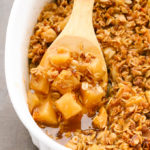 Close-up overhead view of Healthy Apple Crisp in a large white serving dish with a wooden spoon taking a spoonful out.