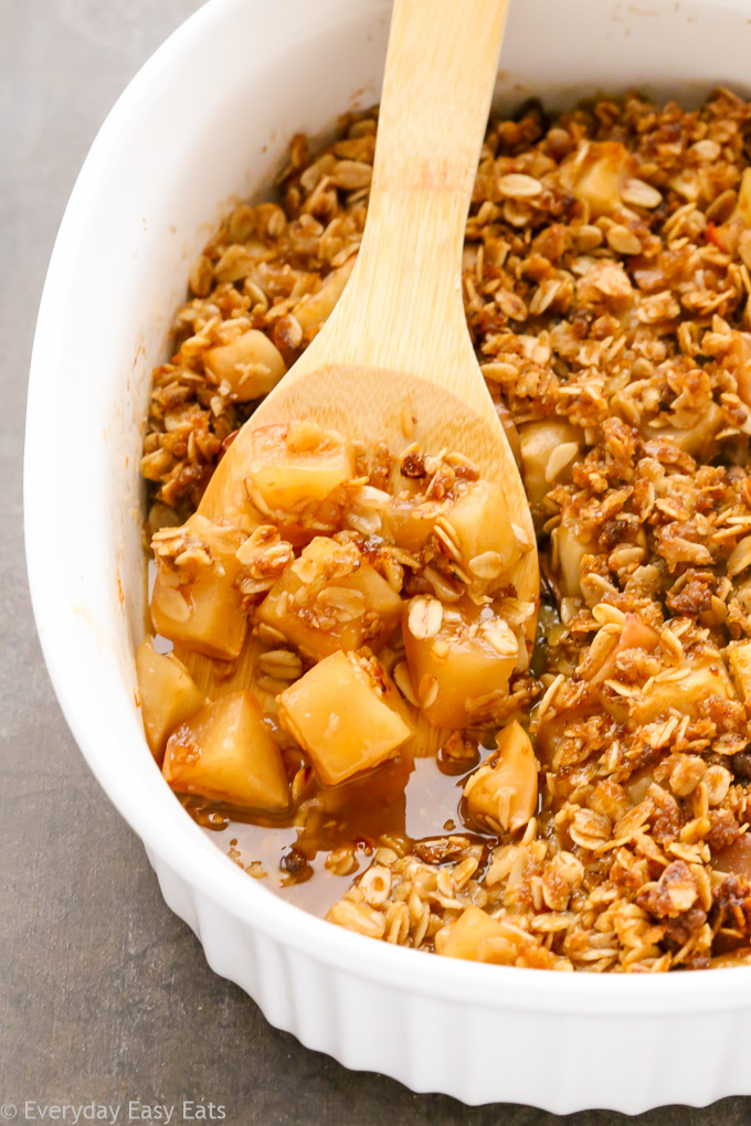 Close-up overhead view of Healthy Apple Crisp in a large white serving dish with a wooden spoon taking a spoonful out.