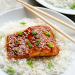 A plate of Baked Honey Sriracha Salmon sitting on a bed of white rice with chopsticks.