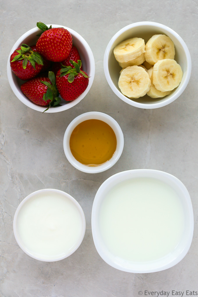 Overhead view of Strawberry Banana Smoothie ingredients in white bowls on a light background.