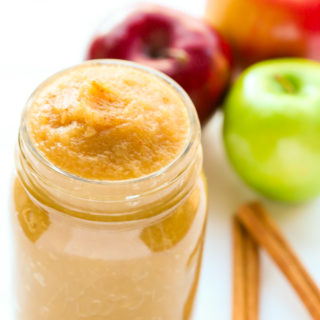 Overhead view of a open jar of Unsweetened Applesauce with apples and cinnamon sticks behind it on a white background.