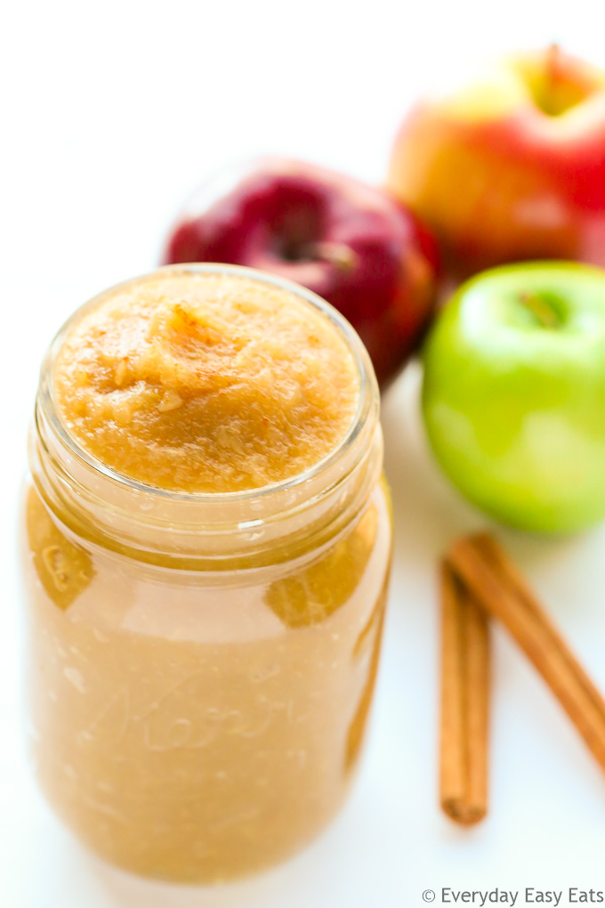 Overhead view of a open jar of Unsweetened Applesauce with apples and cinnamon sticks behind it on a white background.