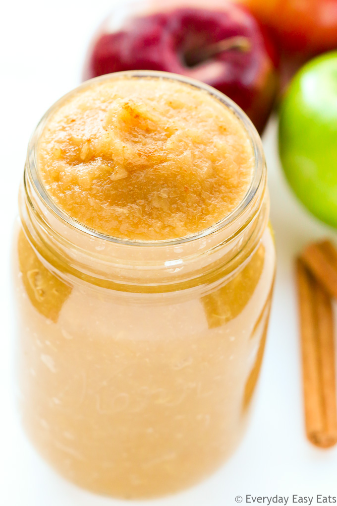 Close-up overhead view of a open jar of Unsweetened Applesauce with apples and cinnamon sticks behind it on a white background.