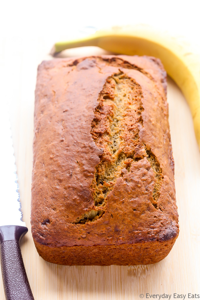Overhead view of a loaf of Moist Banana Bread on a wooden chopping board with a bread knife on the side.