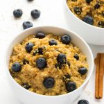 Overhead view of two Blueberry Quinoa Breakfast Bowls on a white background with scattered blueberries and cinnamon sticks.