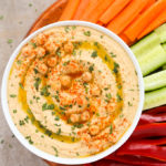 Overhead view of a bowl of Hummus Without Tahini with chopped vegetables on a neutral background.