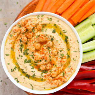 Overhead view of a bowl of Homemade Hummus Without Tahini with chopped vegetables on a neutral background.