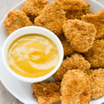 Overhead view of Panko Baked Chicken Nuggets in white plate with a small dipping bowl of honey mustard sauce on the side.