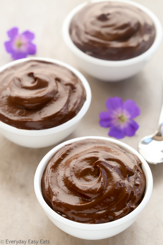 Overhead view of three bowls of Healthy Chocolate Avocado Pudding with purple flowers scattered around.