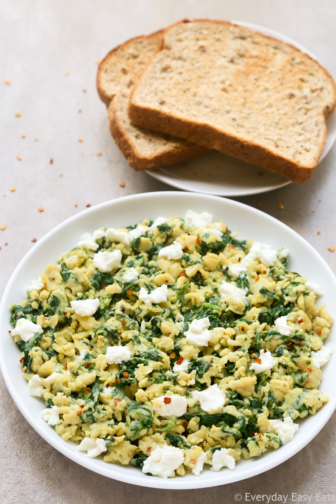 Overhead view of a plate of Scrambled Eggs with Spinach with toast on the side on a neutral-colored surface.