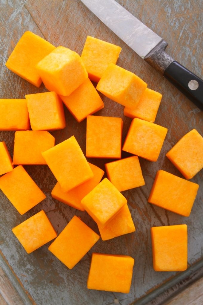 Close-up overhead view of fresh butternut squash diced into cubes on a chopping board with a chef's knife on the side.