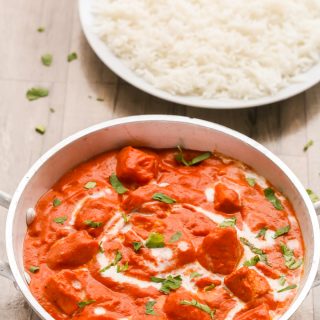Overhead view of Easy Butter Chicken in a silver serving bowl with a plate of white rice on a wooden background.