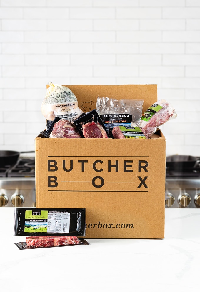 ButcherBox Review: Opened ButcherBox delivery box filled with various frozen meat packages set on a white marble table in a white kitchen.