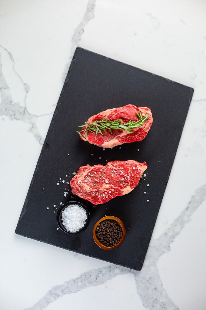 ButcherBox Review: Two raw grass-fed beef steaks with seasonings on a black cutting board placed on a white marble table.