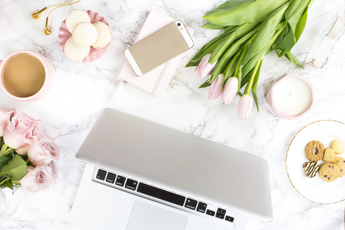 Contact Page for Everyday Easy Eats: Overhead view of a silver laptop, cell phone, cup of coffee, and fresh flowers on a white marble desk.